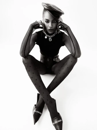 Black and white fashion portrait of an adult woman in patterned tights, a leather beret, a top and Balenciaga footwear in a studio.