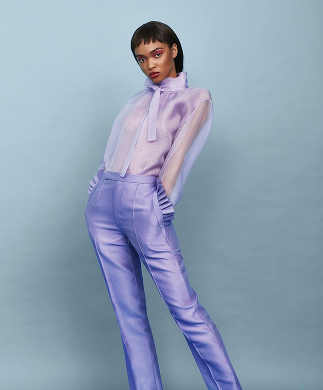 Fashion shot of a female model with dark hair posing in a light purple silk blouse and trousers against a baby blue background.