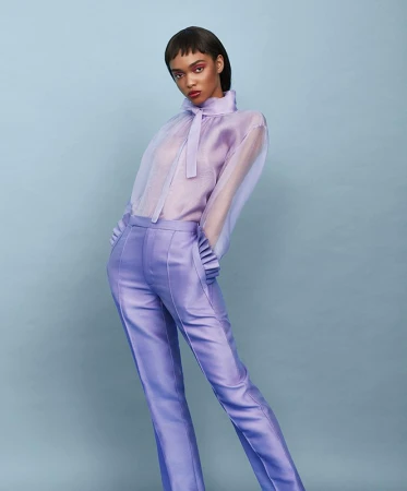 Fashion shot of a female model with dark hair posing in a light purple silk blouse and trousers against a baby blue background.