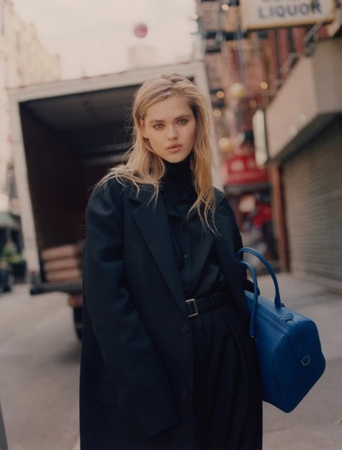 A woman in a black coat and dress poses with a blue bag in an urban street setting with an out-of-focus delivery truck behind her.