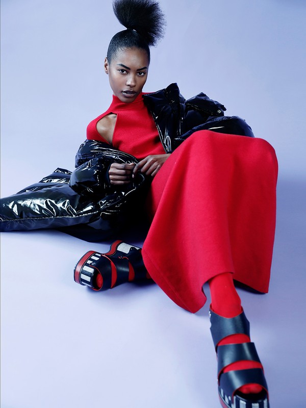 Fashion shot of a woman in a red dress and black puffy coat. The model is seated against a light purple studio backdrop.