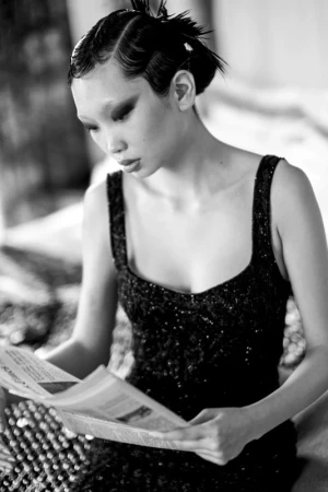 Elegant woman in black dress with intricate hairstyle reads newspaper, creating a vintage editorial moment in a softly lit indoor setting.