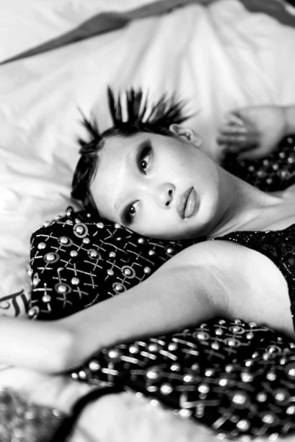 Dramatic black-and-white portrait of a stylish adult female with spiky hair and dramatic eye makeup reclining on a bed.