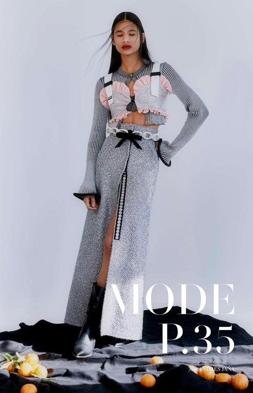 Fashion editorial shot of an adult woman in a stylish sweater crop top and a long slit skirt, posing against a neutral background with oranges.