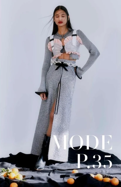 Fashion editorial shot of an adult woman in a stylish sweater crop top and a long slit skirt, posing against a neutral background with oranges.