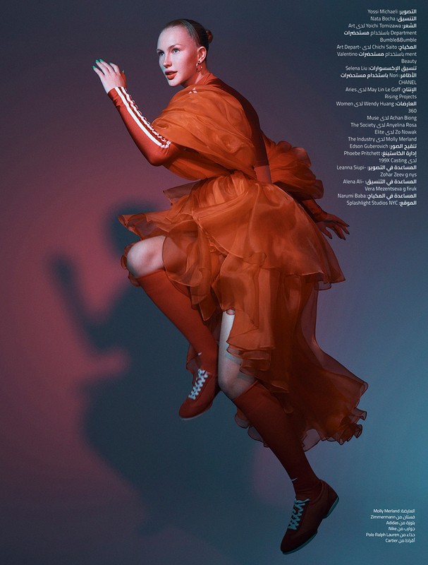 Fashion editorial shot of a slender woman in an orange layered dress and matching shoes, posing in front of a red and blue gradient background.