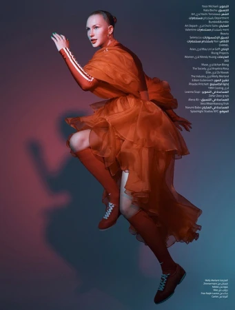 Fashion editorial shot of a slender woman in an orange layered dress and matching shoes, posing in front of a red and blue gradient background.