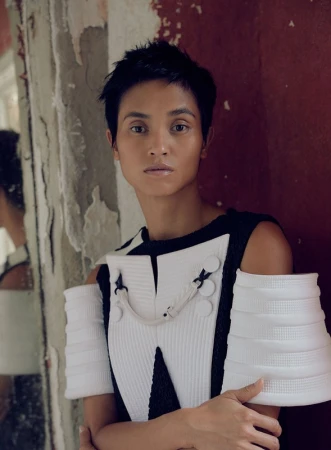 Portrait of a model with short dark hair wearing a structured black and white top with sculptural pleated sleeves against a rustic wall
