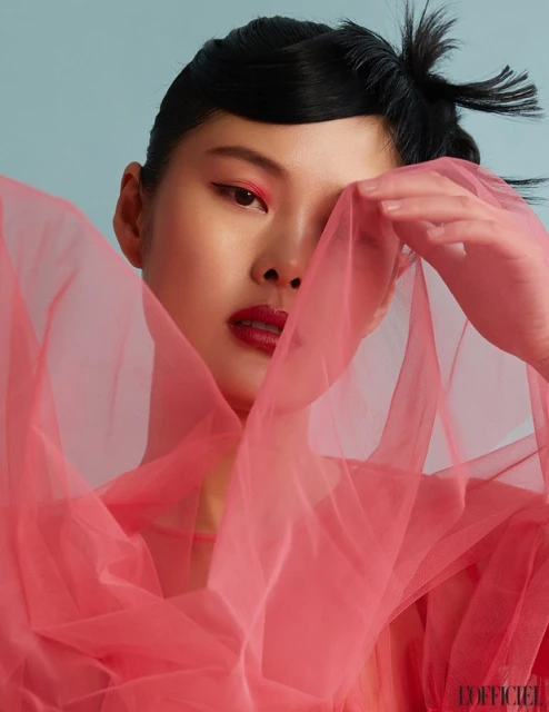 Asian woman with dark hair, wearing vivid red makeup and a sheer red shawl, poses against a light blue backdrop in a close-up shot.