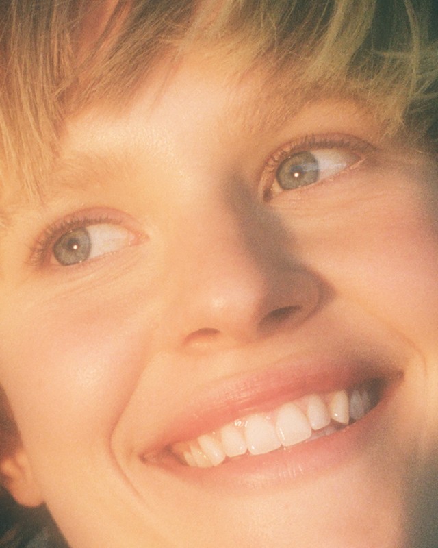 Close-up portrait of a young adult woman with fair skin and blonde hair smiling broadly, showing her white teeth.