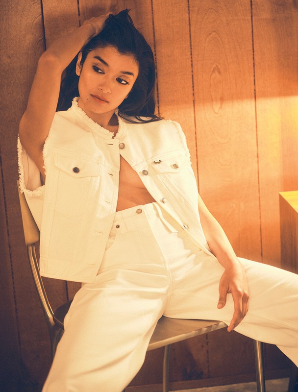 Brunette woman posing while sitting on a metal chair, wearing a white denim vest and pants in front of a wood paneled wall.