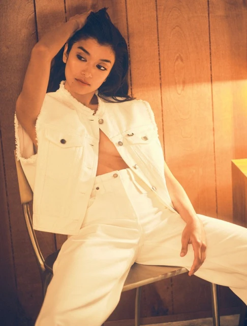 Brunette woman posing while sitting on a metal chair, wearing a white denim vest and pants in front of a wood paneled wall.