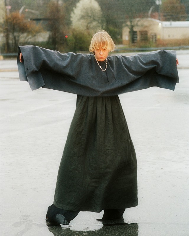 A person with short blonde hair stands in a wet parking lot wearing a draped gray top and black wide-legged pants with arms outstretched.