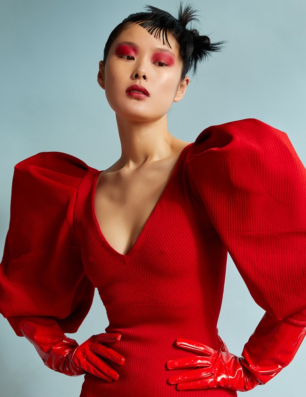 Asian woman in red dress and gloves strikes a pose against a pale blue backdrop, showcasing beauty and fashion.