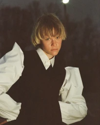 Portrait of a model with short blonde hair wearing a black vest and white puffy-sleeved shirt outdoors at night.
