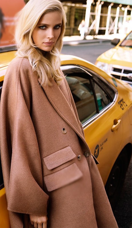 Fashion editorial shot of a blonde woman wearing a brown coat, standing next to a New York City yellow taxi cab on a city street.