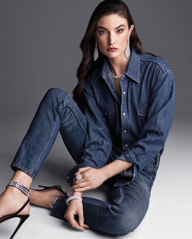 Brunette woman poses in denim shirt and jeans while showcasing diamond jewelry including earrings, necklace, bracelet, and ring.