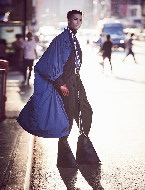 Asian man in high-fashion black bell-bottoms and blue coat poses on city street with blurred background of cars and pedestrians.