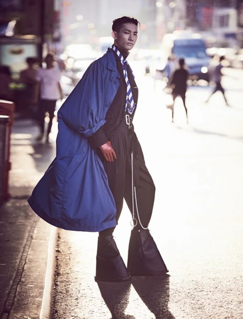 Asian man in high-fashion black bell-bottoms and blue coat poses on city street with blurred background of cars and pedestrians.