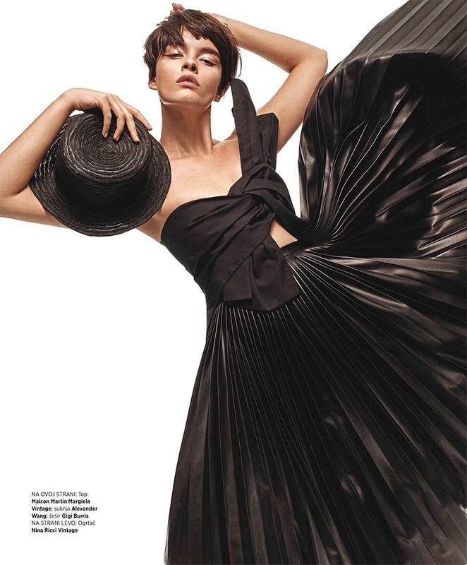 Fashion editorial shot of a brunette woman in a black one-shoulder dress holding a black straw hat, set against a stark white background.
