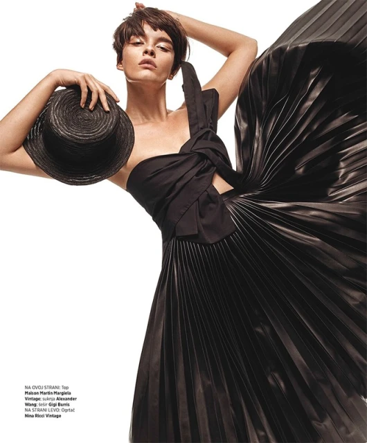 Fashion editorial shot of a brunette woman in a black one-shoulder dress holding a black straw hat, set against a stark white background.