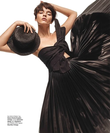 Fashion editorial shot of a brunette woman in a black one-shoulder dress holding a black straw hat, set against a stark white background.
