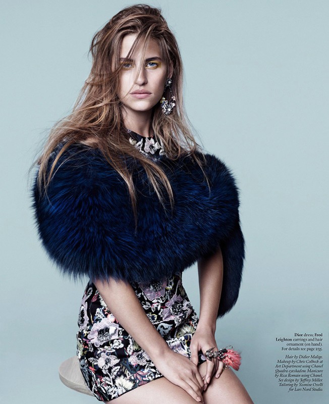 A model poses in a studio wearing a floral dress with a blue fur stole, statement earrings, and a ring. Light blue background.