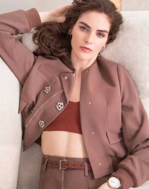 Woman poses on a couch wearing a jacket, top, and trousers in shades of brown and rust. She has brown hair and fair skin.