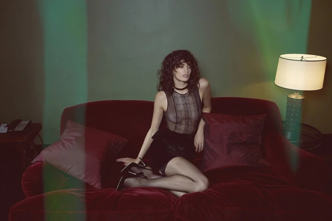 A woman with curly hair poses on a red velvet sofa wearing a black lace bodysuit, matching short skirt, stockings, and heels in an indoor setting.