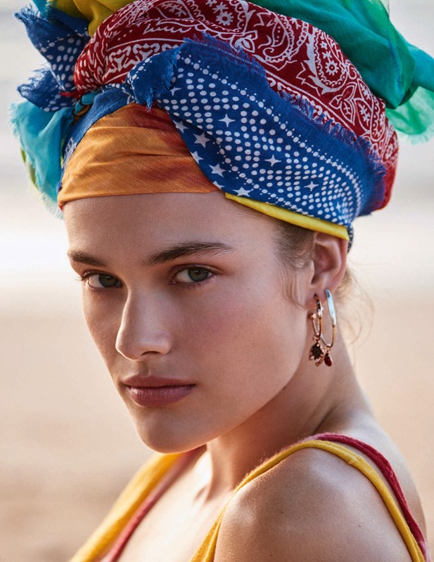 Close-up shot of a woman with a colorful scarf wrapped around her head, wearing gold hoop earrings, with a blurred outdoor background.