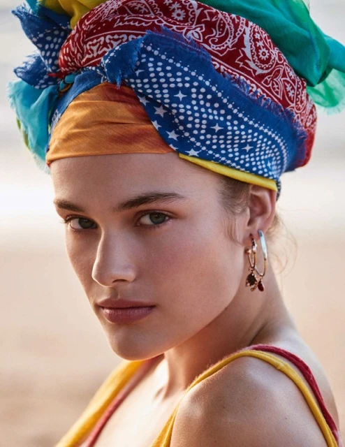 Close-up shot of a woman with a colorful scarf wrapped around her head, wearing gold hoop earrings, with a blurred outdoor background.
