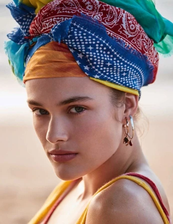 Close-up shot of a woman with a colorful scarf wrapped around her head, wearing gold hoop earrings, with a blurred outdoor background.