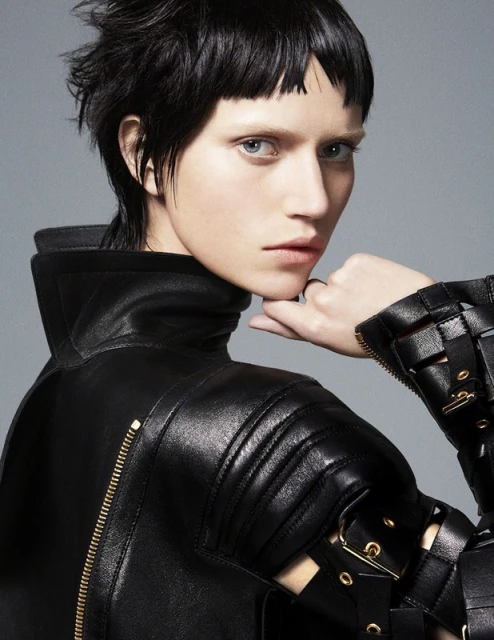 Close-up studio shot of an adult woman with a short, dark hairstyle wearing a high-collared black leather jacket and metal accents.