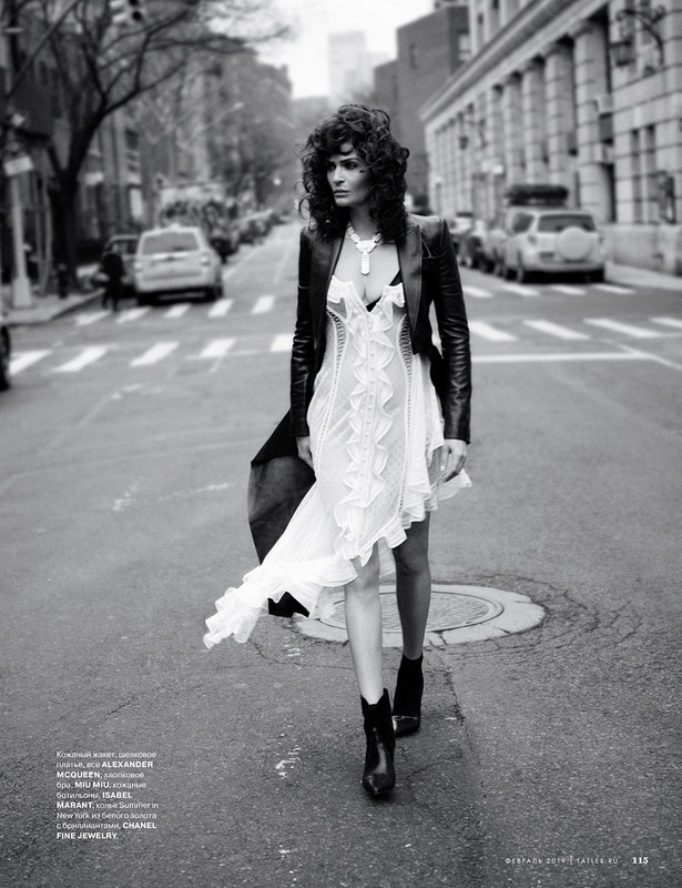 Fashionable woman in dress, leather jacket walking on street in black-and-white, featuring cars, buildings, and urban setting.