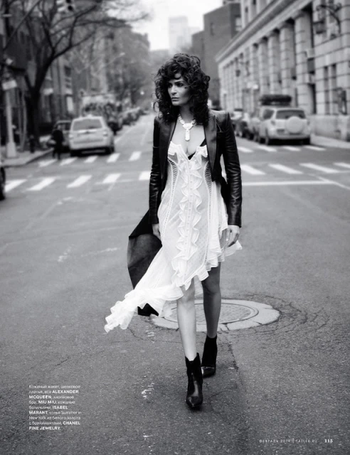 Fashionable woman in dress, leather jacket walking on street in black-and-white, featuring cars, buildings, and urban setting.