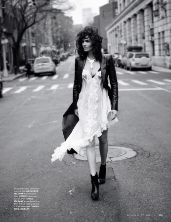 Fashionable woman in dress, leather jacket walking on street in black-and-white, featuring cars, buildings, and urban setting.