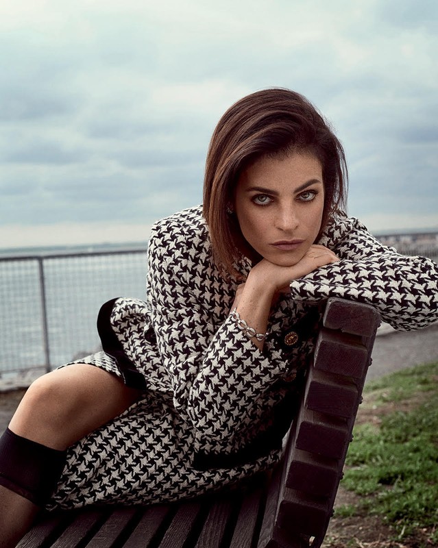 Woman in houndstooth blazer and skirt sits on a bench in an urban park, posing and looking at the camera with a serious expression.
