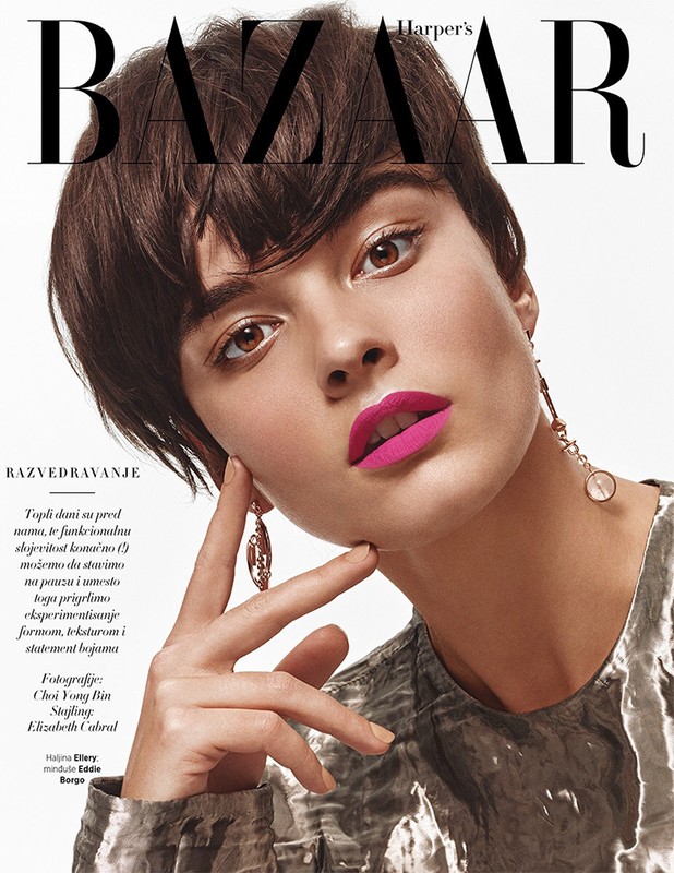 Close-up beauty portrait of a woman with short dark hair, vibrant pink lipstick, and dangling earrings against a white background.