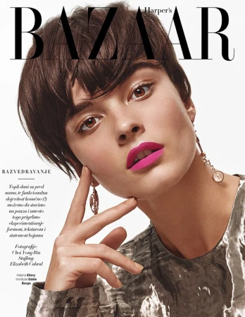 Close-up beauty portrait of a woman with short dark hair, vibrant pink lipstick, and dangling earrings against a white background.