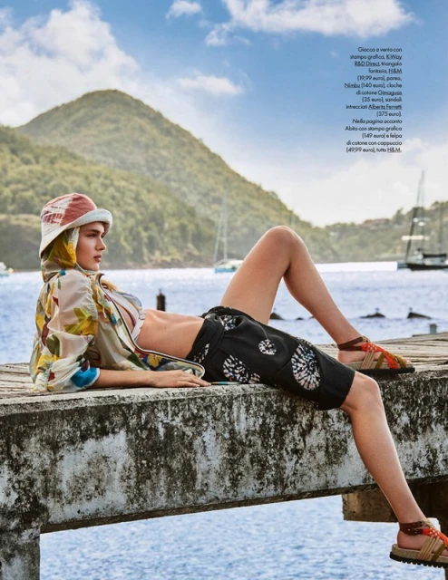 A woman in a floral jacket and shorts reclines on a pier by the ocean with lush green mountains and boats in the background under a cloudy sky.
