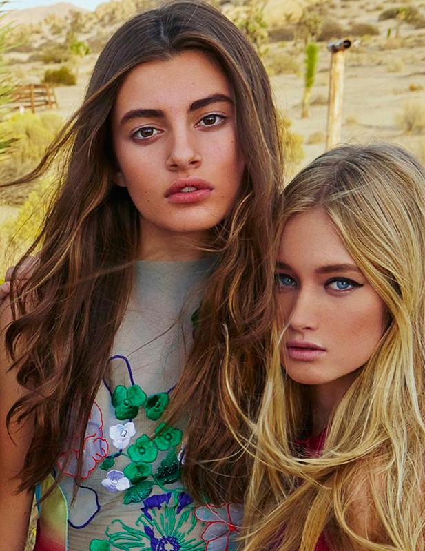 Two young women pose closely together in a sunlit outdoor setting. They both have long hair and are wearing sheer tops with floral details.