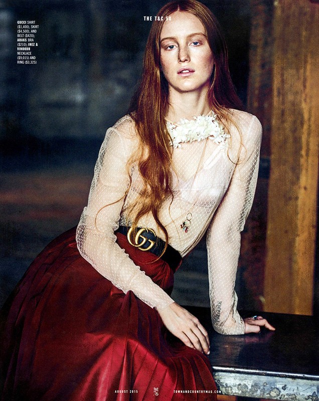 A red-haired model poses in an indoor fashion shot, wearing a sheer blouse, a red skirt, and Gucci belt. The setting features rustic columns.