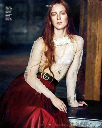 A red-haired model poses in an indoor fashion shot, wearing a sheer blouse, a red skirt, and Gucci belt. The setting features rustic columns.