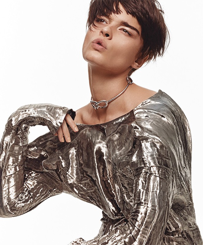 Brunette woman in shiny, metallic silver top and modern metal necklace poses in a studio with bright, neutral lighting on white background.