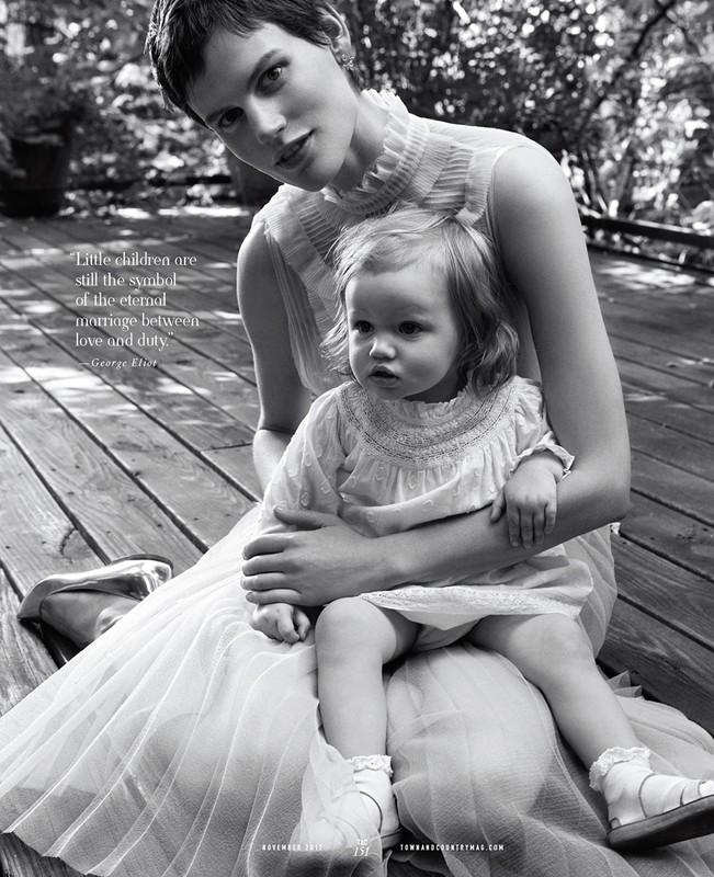Monochrome portrait of a woman in tulle dress holding a toddler in lace dress, both seated on a wood deck in a leafy outdoor setting.