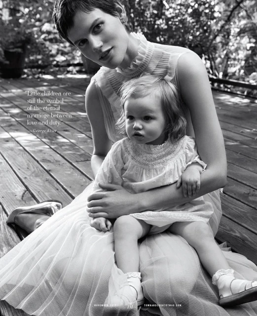 Monochrome portrait of a woman in tulle dress holding a toddler in lace dress, both seated on a wood deck in a leafy outdoor setting.