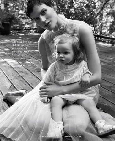 Monochrome portrait of a woman in tulle dress holding a toddler in lace dress, both seated on a wood deck in a leafy outdoor setting.