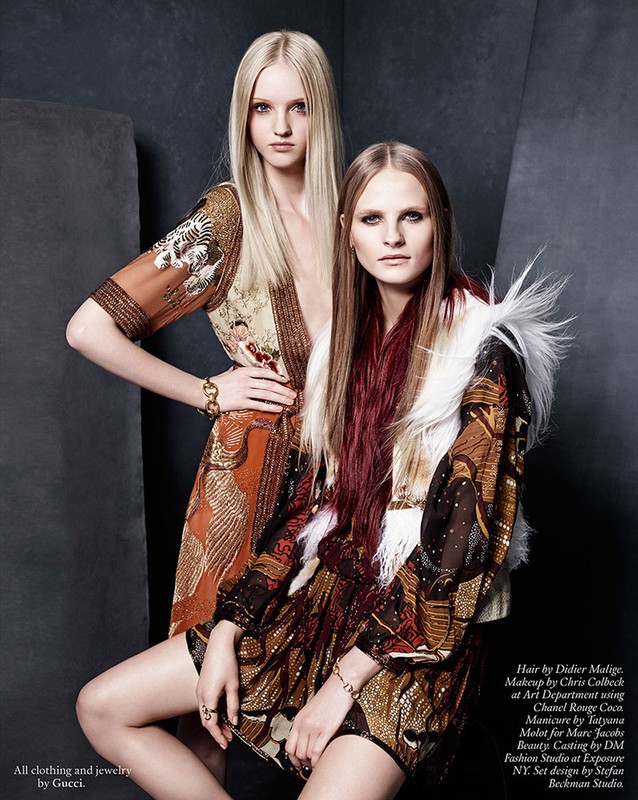 Two fashion models pose in a studio wearing patterned mini-dresses and bracelets. The blond model has her hand on the shoulder of the seated model.
