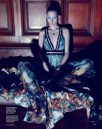 Model with vibrant blue eye makeup wearing a patterned long gown seated against wood panels
