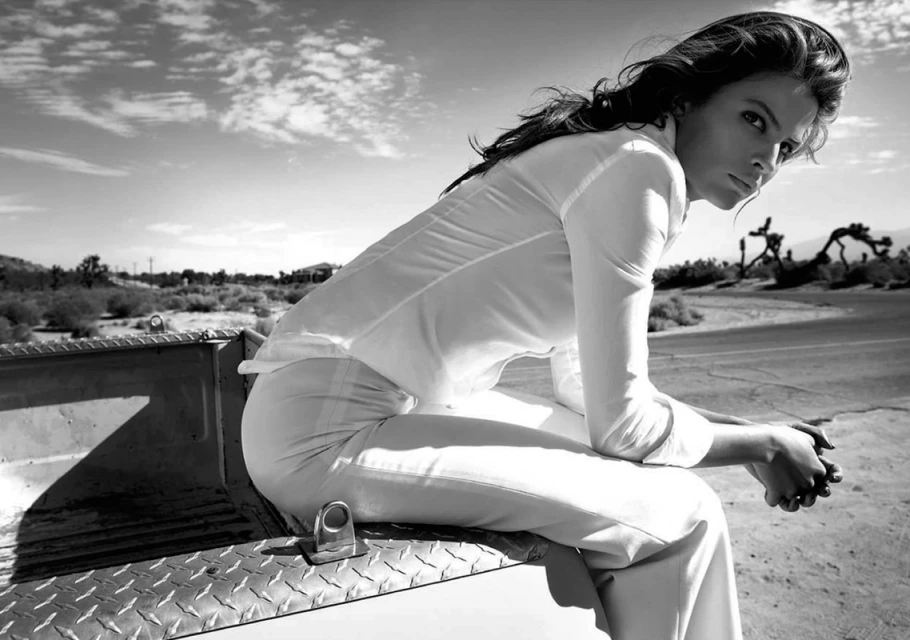 Black and white outdoor portrait of a woman with long dark hair, wearing a white shirt and pants, sitting in the back of a truck.